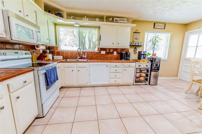 Bright and spacious kitchen with white cabinetry and appliances, featuring warm tile countertops and backsplash. Large windows bring in natural light, and there's ample floor space for movement. Ideal for cooking and entertaining. | Image 15