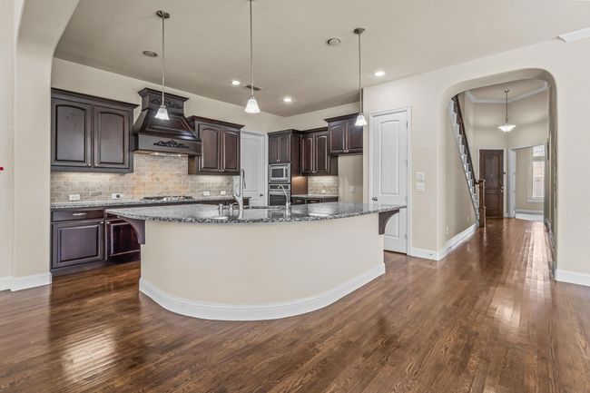 Kitchen with custom exhaust hood, dark brown cabinetry, an island with sink, decorative backsplash, and dark stone countertops | Image 10