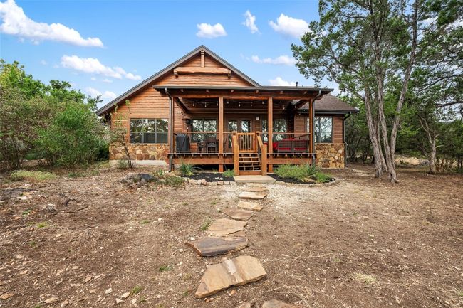 The back of the home showcases its rustic charm, with natural wood siding and stone accents that blend seamlessly with the landscape. A welcoming back deck, complete with room for seating and dining overlooks the tranquil surroundings, accessible by a stone path that adds to the natural feel of the property. | Image 22