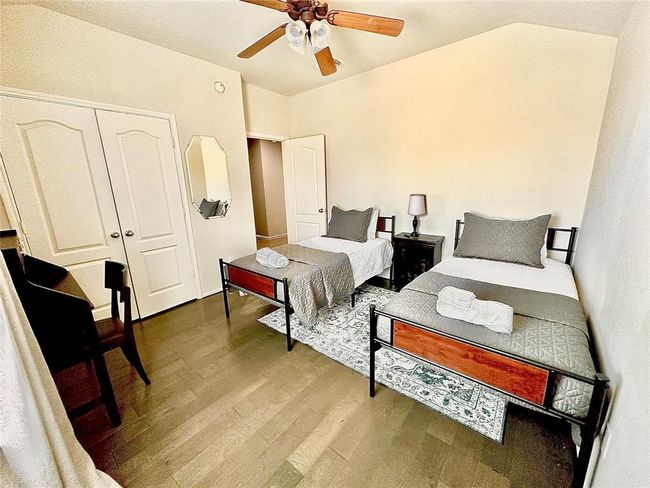 This cozy bedroom features two twin beds with matching gray bedding, a ceiling fan, and hardwood floors. There’s a small desk and chair, a bedside table with a lamp, and a closet with double doors, perfect for guests or children. | Image 14