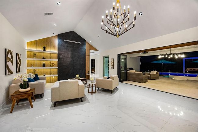 This striking living area impresses with its soaring vaulted ceiling, dramatic black stone accent wall, and custom built-in shelving with warm wood tones and integrated lighting. Large-format polished tile floors enhance the sense of space and luxury, while retractable glass walls blur the line between indoor and outdoor living, opening to a covered patio and an illuminated pool oasis beyond. A perfect blend of contemporary design and functional elegance. | Image 8
