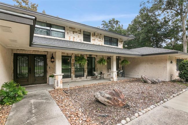 This photo shows a two-story home with a stone exterior and a covered front porch. The entrance features decorative wrought iron doors, and the porch is adorned with hanging plants. The yard is landscaped with stones and mature trees, offering a welcoming curb appeal. | Image 7