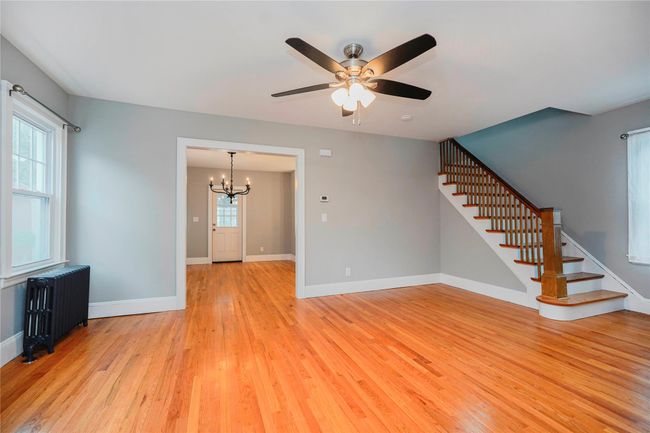 Unfurnished living room with ceiling fan with notable chandelier, stairway, radiator, and light wood finished floors | Image 7