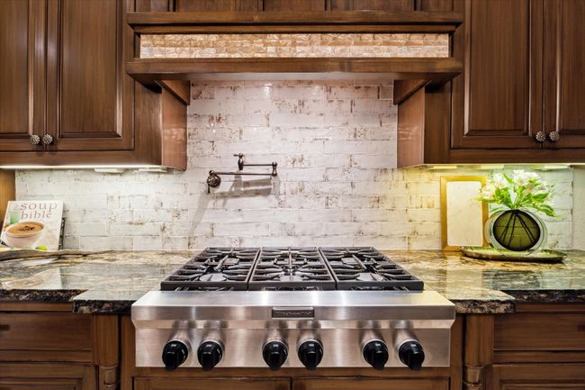 A culinary centerpiece, this professional-grade KitchenAid 6–burner gas cooktop is set against a hand-glazed brick backsplash and topped with a custom wood range hood featuring an accent of iridescent mother of pearl tile. The richly veined granite countertops add warmth and sophistication, while under-cabinet lighting and a convenient pot filler elevate both form and function. Every detail is thoughtfully curated for style and performance. | Image 18