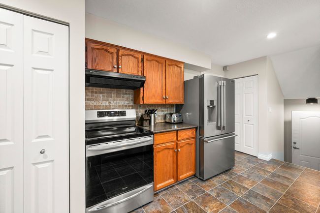 Kitchen featuring dark countertops, brown cabinets, decorative backsplash, range hood, and appliances with stainless steel finishes | Image 11