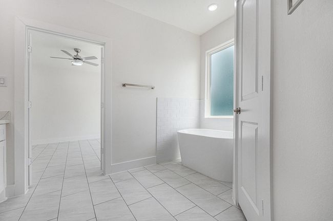 This photo showcases a bright, modern bathroom with a standalone bathtub, large frosted window for privacy, and sleek tiled flooring. The space connects to an adjoining room with a ceiling fan, offering an open and airy feel. | Image 24