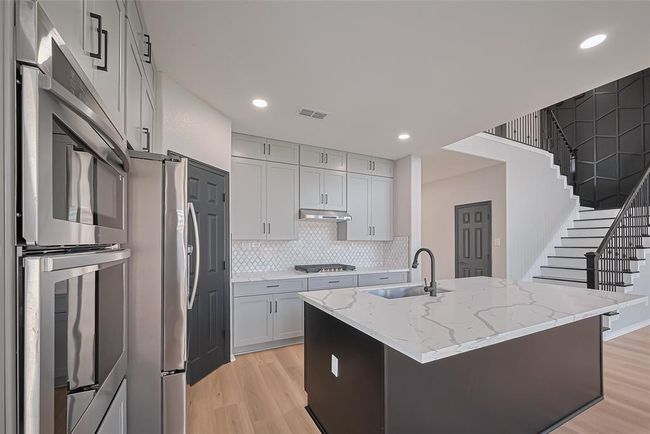 Modern kitchen featuring stainless steel appliances, sleek cabinetry, and a large island with a marble countertop. The open layout includes adjacent stairway access, enhancing the spacious feel. | Image 12