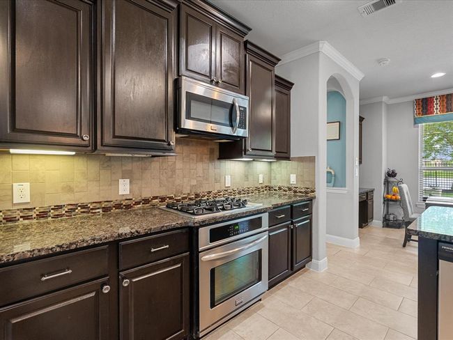 This spacious kitchen and dining area features modern appliances, a large island with pendant lighting, dark cabinetry, and a tile floor. It opens to a bright living space with large windows and a door leading to the outdoor area, perfect for entertaining and family gatherings. | Image 9