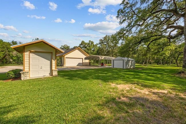 Cluster of functional outbuildings including the wellhouse, two car garage, covered carport and additional metal storage building—all situated on lush, open lawn under mature oaks. | Image 33