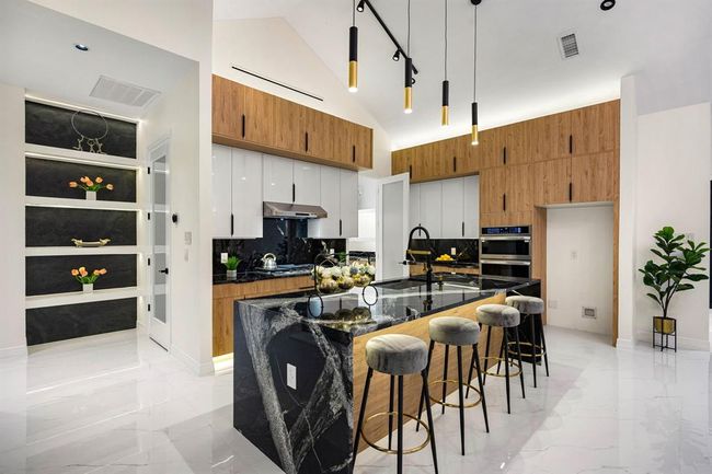 Step into this stunning modern kitchen featuring sleek high-gloss soft close cabinetry, warm wood finishes, and elegant black marble countertops. The spacious island offers ample workspace, perfectly complemented by dramatic pendant lighting and a vaulted ceiling that enhances the open, airy feel. High-end built-in appliances and under-cabinet lighting add both functionality and style, while the polished marble floors tie the space together with a luxurious touch. | Image 11
