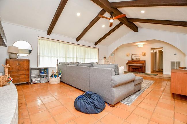 This inviting living room features a high ceiling with exposed wooden beams, terracotta tile flooring, and abundant natural light from a large window. A cozy gray sectional, warm wood accents, and a ceiling fan add comfort and style. | Image 14
