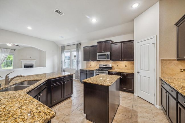 This kitchen features dark wood cabinetry, granite countertops, a central island, and stainless steel appliances. It offers a spacious layout with tiled flooring and recessed lighting, perfect for modern living. | Image 13