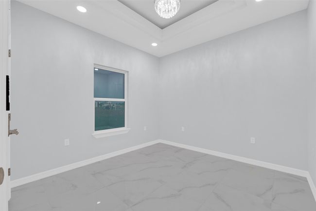This room features a modern design with light gray walls, polished marble tile flooring, and a recessed ceiling with a chandelier. A large window provides natural light, creating an inviting atmosphere. Perfect for a bedroom or home office. | Image 25