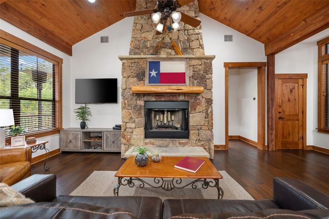 The centerpiece of this cozy living room is a stone fireplace adorned with a Texas flag, blending rustic charm with local pride. Natural wood finishes and large windows bring warmth and light to the space. | Image 7