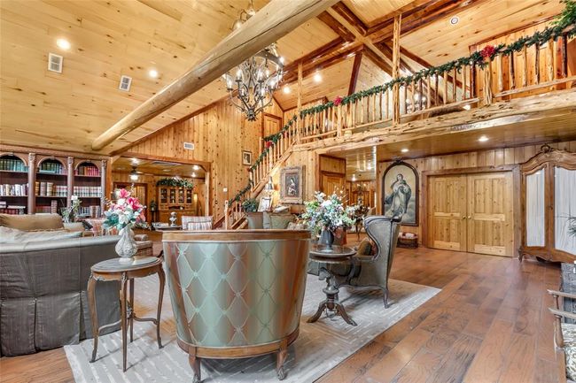 This spacious living area features rustic wood paneling and ceilings, a grand staircase with a loft, and elegant wood beams. The room is tastefully decorated with floral arrangements, a large chandelier, and built-in bookshelves, offering a cozy yet sophisticated atmosphere. | Image 26