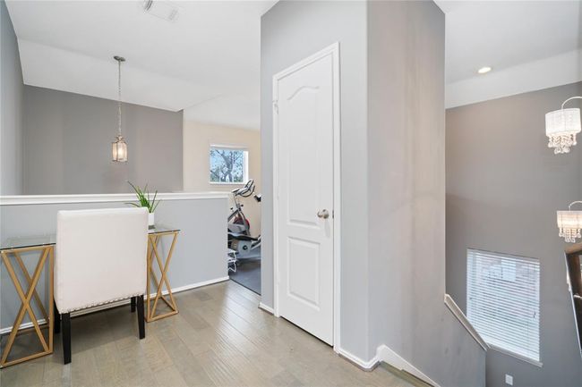 This image showcases a bright, modern upstairs hallway with wooden flooring and a neutral color palette. It features a small desk area under a hanging light fixture, adjacent to a room with exercise equipment. The staircase is visible with elegant lighting and a window providing natural light. | Image 40