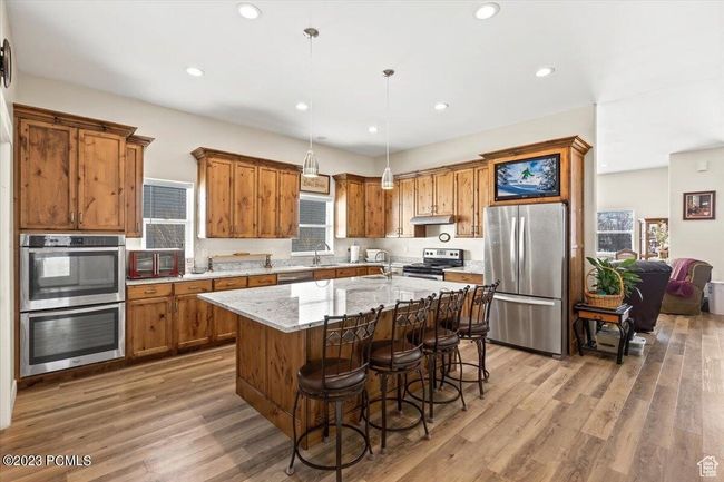 Kitchen featuring stainless steel appliances, brown cabinetry, wood finished floors, an island with sink, and under cabinet range hood | Image 6