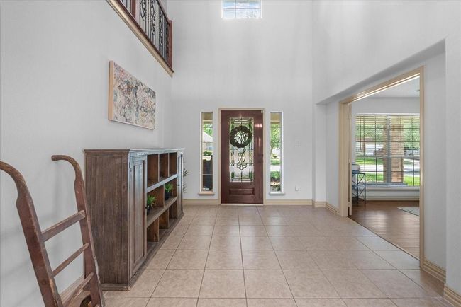 This entryway features a high ceiling with natural light, a decorative wooden front door, tile flooring, and an open view into a bright room with large windows. | Image 5