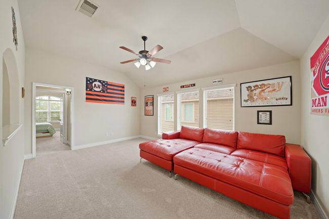 This photo showcases a bright, spacious room with a high ceiling. The space is carpeted and well-lit by multiple windows. A ceiling fan is centrally located, and there is an open doorway leading to a principal bedroom. | Image 26