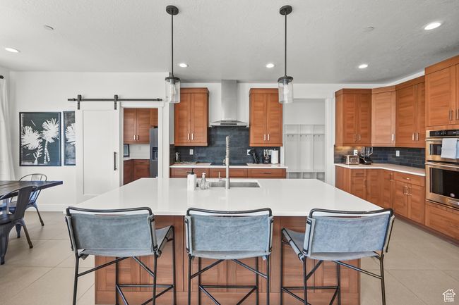 Kitchen with wall chimney exhaust hood, brown cabinets, double oven, backsplash, and light countertops | Image 12