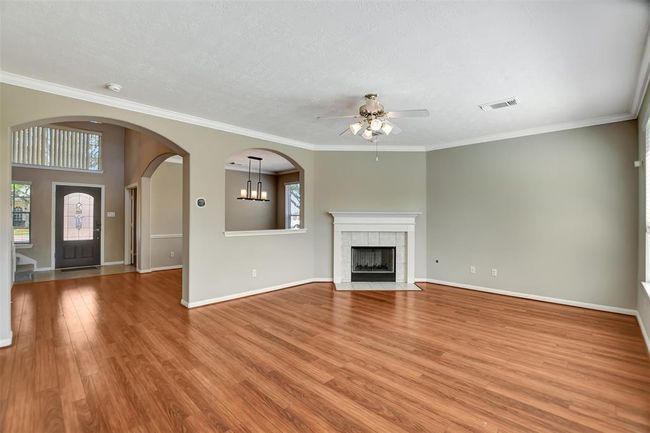 This spacious living room features warm wood flooring, a cozy fireplace, and elegant crown molding. An open archway leads to the dining area, enhancing the home's open feel. A ceiling fan adds comfort, while large windows provide natural light. | Image 13