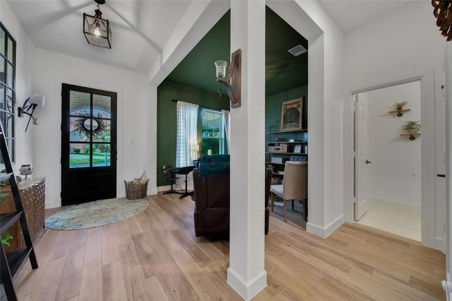 This entryway features a welcoming space with light wood flooring, a stylish black front door, and a cozy sitting area in an alcove with green walls. Natural light fills the room through large windows, creating a bright and inviting atmosphere. | Image 13