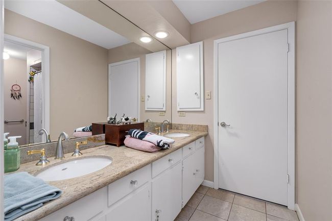This secondary bathroom features a spacious dual-sink vanity with a granite countertop and ample cabinet storage. A large mirror enhances the brightness of the space, complemented by recessed lighting. The neutral color palette and tiled flooring create a clean and inviting atmosphere. A separate bathing area with a tiled shower adds functionality and privacy. | Image 30