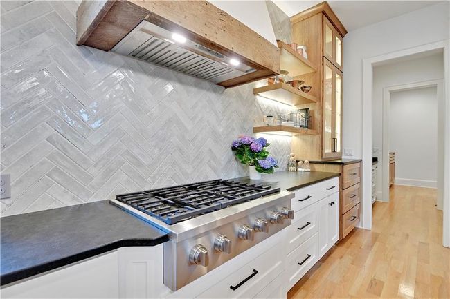 Kitchen featuring dark countertops, stainless steel gas stovetop, glass insert cabinets, custom range hood, and backsplash | Image 21