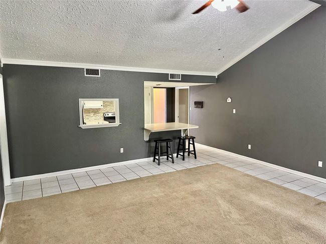 This room features a spacious open layout with a mix of carpet and tile flooring. It has a vaulted ceiling with a ceiling fan, a breakfast bar with stools, and a pass-through window to the kitchen. The walls are painted in a modern gray tone. | Image 5