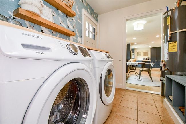 This photo showcases a well-lit laundry room featuring a modern washer and dryer set. The space is accented with stylish wallpaper and open wooden shelves, leading into a bright kitchen area. | Image 41