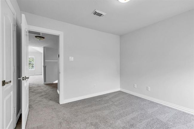 This photo shows a small, carpeted room with neutral walls, ideal as a bedroom or office. It features a door leading to a hallway with additional natural light from a window, creating a bright and open feel. | Image 39