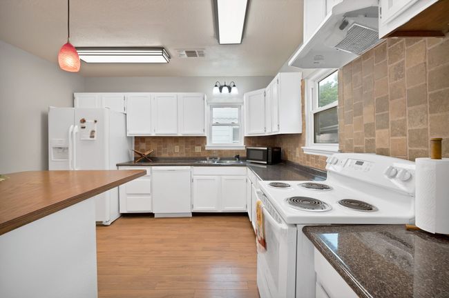 Kitchen featuring dark granite countertops, backsplash, bar, under cabinet range hood, white appliances, and white cabinetry, ventilation hood, white appliances, and white cabinetry | Image 14