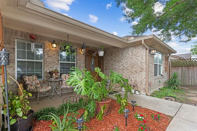 This is a cozy, well-maintained front porch featuring brick exterior walls, two large windows, and a covered sitting area with comfortable furniture. The space is accented by lush potted plants, decorative outdoor lighting, and a small landscaped garden area with vibrant flowers, creating a welcoming entrance to the home. | Image 7