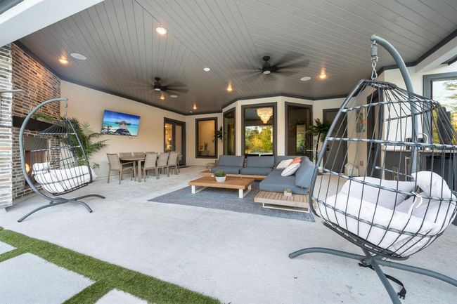 A spacious, modern outdoor living area with a covered patio. It features a cozy seating arrangement, dining table, hanging chairs, a fireplace, and a mounted TV. The area is well-lit with ceiling fans and recessed lighting, providing a comfortable and stylish space for relaxation and entertainment. | Image 41