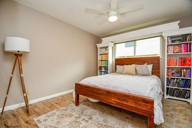 Cozy bedroom with warm wood flooring, a stylish wooden bed, and built-in bookshelves flanking a large window. The neutral walls and modern ceiling fan create a serene atmosphere, complemented by a decorative floor lamp and plush area rug. | Image 35