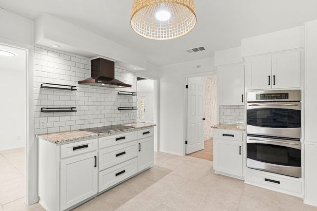 Kitchen featuring wall chimney exhaust hood, double oven, black electric stovetop, backsplash, and white cabinetry | Image 12