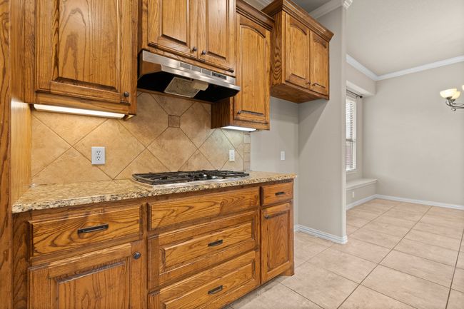 Kitchen with under cabinet range hood, stainless steel gas cooktop, crown molding, tasteful backsplash, and brown cabinetry | Image 17