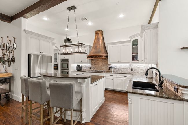 Kitchen featuring appliances with stainless steel finishes, solid wood floors, crisp white custom built cabinets, granite countertops, a decorative stone backsplash, a custom copper vent hood, breakfast bar and decorative lighting. | Image 11
