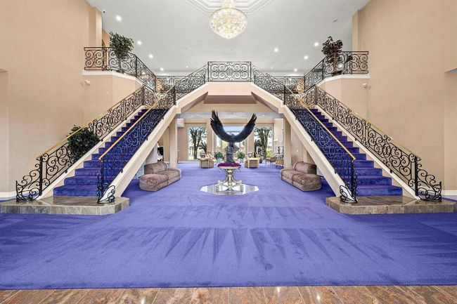 This is a grand foyer with dual sweeping staircases, ornate wrought-iron railings, and a bold purple carpet, leading to an elegant second-floor landing. The space is open and airy, featuring a high ceiling with a chandelier, enhancing the luxurious feel of the home. | Image 5