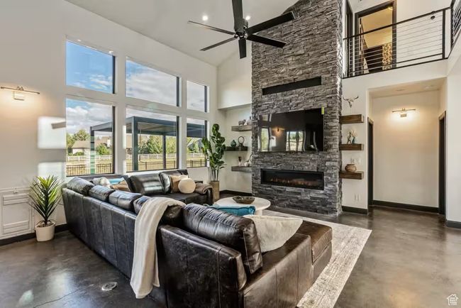 Living area with finished concrete flooring, a high ceiling, a ceiling fan, a stone fireplace, and baseboards | Image 15