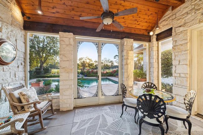 This photo showcases a charming patio with stone walls and a wooden ceiling, featuring a ceiling fan and comfortable seating. It overlooks a lush garden and a swimming pool, offering a serene view and inviting outdoor space. | Image 30