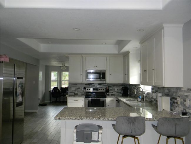 Modern kitchen with granite countertops and stainless steel appliances. It features ample cabinet space, a tiled backsplash, and a breakfast bar with seating. The open layout is complemented by natural light and recessed lighting. | Image 8