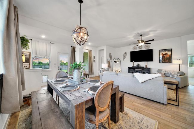 This photo showcases a bright, open-concept living and dining area with modern decor. The space features a cozy living room with a ceiling fan, a wall-mounted TV, and a comfortable sofa. Adjacent is a dining area with a rustic wooden table set for four. Large windows and glass doors allow for ample natural light, creating an inviting atmosphere. | Image 24