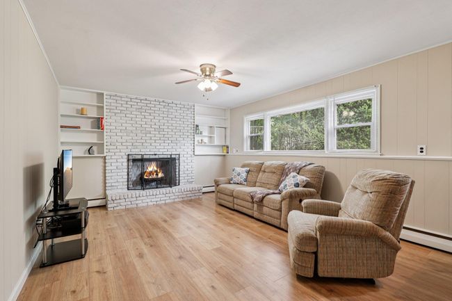 Living room with a ceiling fan, baseboard heating, wood finished floors, a brick fireplace, and crown molding | Image 23