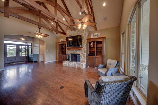 Spacious living room featuring vaulted wooden beams, a cozy stone fireplace, and abundant natural light from large windows. Perfect for relaxation and entertaining. | Image 7