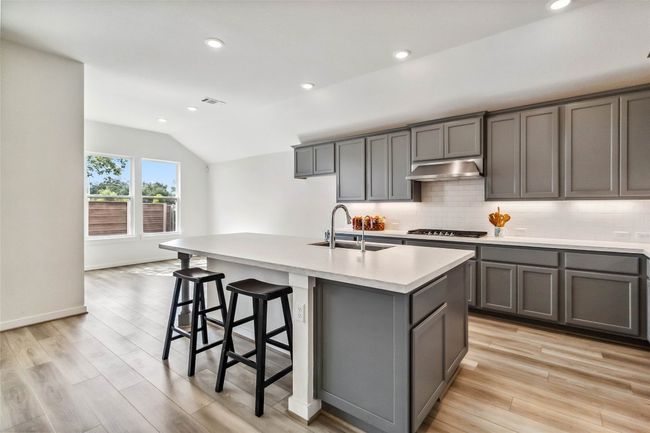 Kitchen with gray cabinets, lofted ceiling, backsplash, a center island with sink, and range hood | Image 12