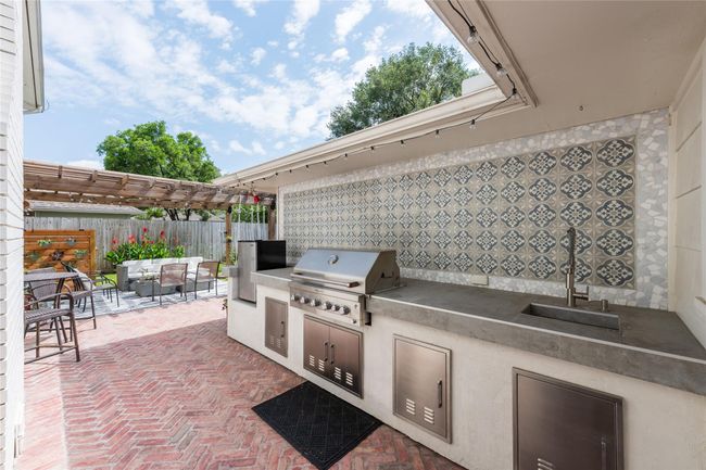 A gorgeous outdoor kitchen with a built-in grill, sink, and patterned tile backsplash. The area features a pergola-covered patio with seating, perfect for entertaining, and a neat brick floor. Lush greenery and a privacy fence complete the inviting backyard space. | Image 4