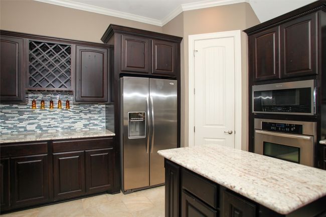 Kitchen featuring appliances with stainless steel finishes, crown molding, dark brown cabinetry, backsplash, and light stone countertops | Image 7
