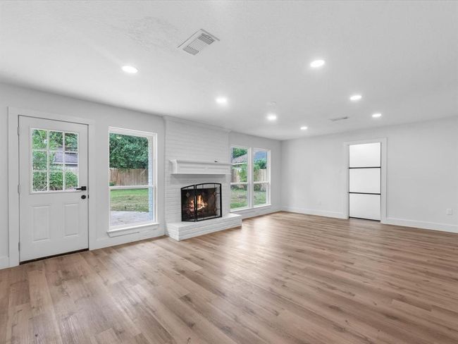 This spacious living room features a cozy fireplace, elegant wood flooring, and ample natural light from large windows. The room offers a bright, airy feel with recessed lighting and a view of the backyard through a glass door. | Image 9