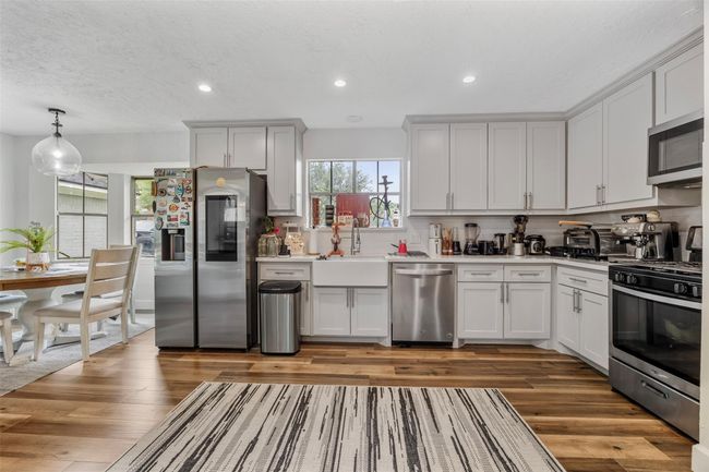 This modern kitchen features sleek white cabinetry, stainless steel appliances, and a farmhouse sink. The open layout connects to a cozy dining area, and natural light fills the space through a large window. | Image 6